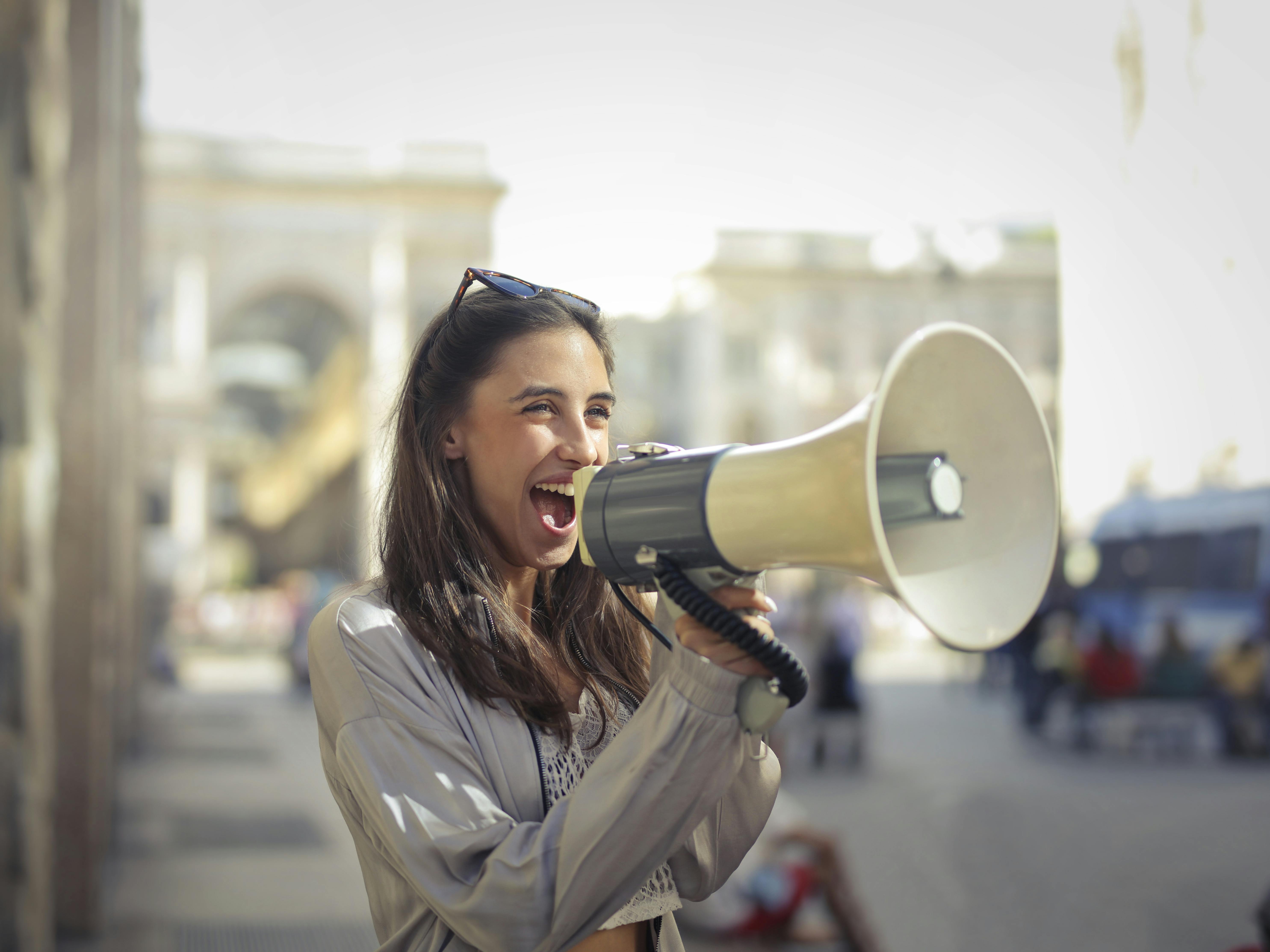 Image of a young women standing in an urban area holding a loudspeaker and speaking into it while smiling.