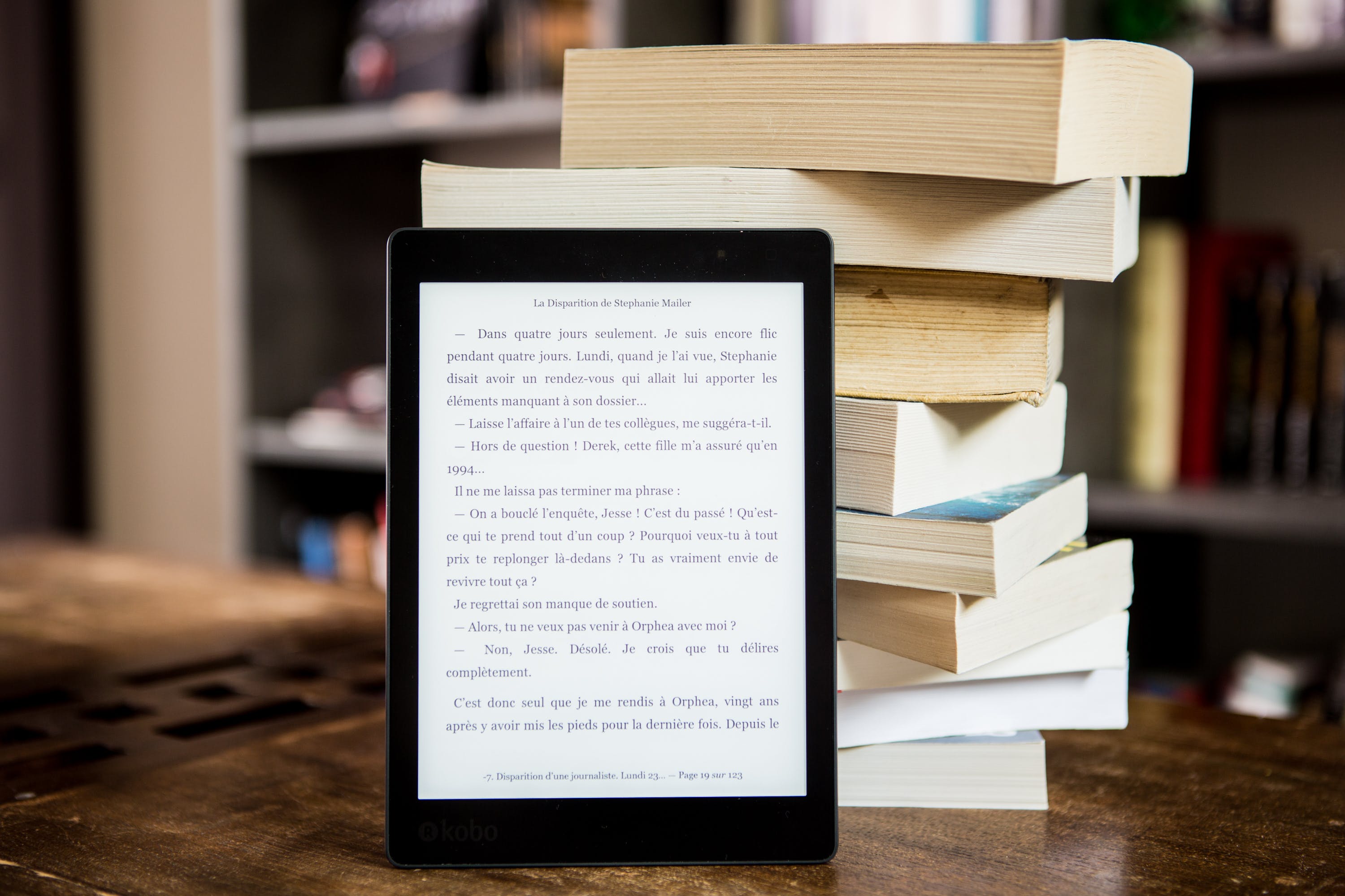 Image of an e-reader leaning against a stack of books on a table in a library.